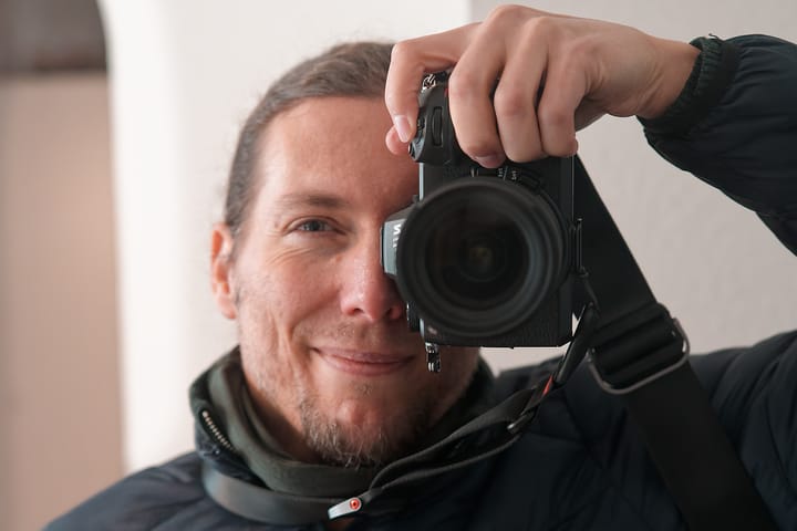 A person holding a mirrorless camera up to their face, photographing themselves indoors. One eye is visible next to the camera body, with a soft, neutral background and natural light.